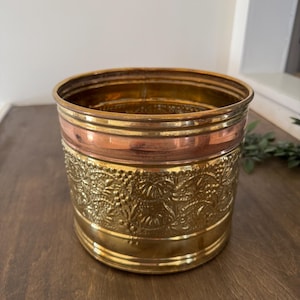 May include: A decorative metal planter with a detailed floral pattern. The planter features bands of gold, copper, and brass. The gold band has an embossed floral design. The planter sits on a wooden surface.
