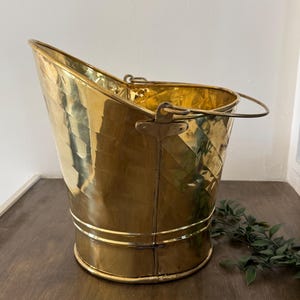 May include: A vintage brass coal bucket with a handle. The bucket has a distinctive shape with a sloped front and a rounded back. The brass has a polished finish, reflecting light. The bucket is sitting on a dark wooden surface.