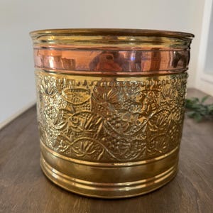 May include: A cylindrical metal planter with a decorative floral pattern. The planter has a copper band at the top and a brass base. The body of the planter is a detailed, embossed gold color. The planter sits on a wooden surface.