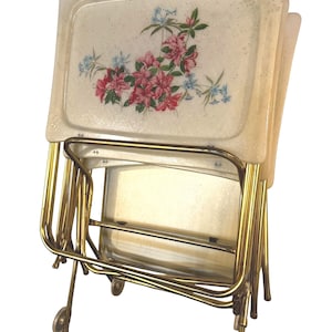 May include: A set of four folding snack trays with gold-tone metal legs and rectangular off-white surfaces featuring a speckled texture and a floral print of pink and blue flowers. The trays are stacked together in a folded, upright position.