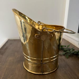May include: A vintage brass bucket with a handle. The bucket has a rounded base and a flared top with a handle. The brass has a polished finish, reflecting the light. The bucket is likely used for coal or other household items.