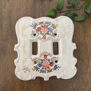 Peut inclure: Plaque de recouvrement d'interrupteur double en céramique crème avec motif floral. La plaque a une forme ornée avec une bordure dorée et deux ouvertures rectangulaires. Le motif floral comprend des fleurs rouges, bleues et roses.
