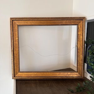 May include: A rectangular, empty picture frame with a gold finish. The frame has a detailed, textured border and is positioned on a dark wooden surface. A thin wire is strung across the empty space, and the frame is against a white wall.