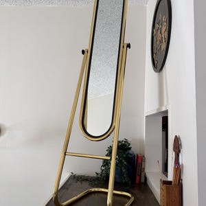 May include: A gold-coloured, full-length mirror with a black frame. The mirror has an oval shape and is supported by a gold metal stand. The stand has a curved base and adjustable height features. The mirror is placed against a white wall.