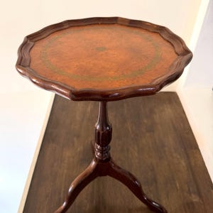 Op de afbeelding: Een kleine, antieke, donkerbruine houten bijzettafel met een geschulpte rand. Het tafelblad is roodbruin met een groen en goudkleurig cirkelvormig ontwerp. De tafel is 71 cm hoog en heeft een driepotige basis.