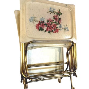 May include: A vintage serving trolley with two cream-coloured trays. The trays feature a floral design with pink and blue flowers. The trolley has a gold-coloured metal frame and small wheels for easy movement.