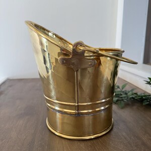 Pode incluir: Um balde de latão dourado com uma alça. O balde tem uma base arredondada e um topo inclinado. O latão é polido e reflete a luz. A alça é fixada com um suporte decorativo. Projetado para transportar carvão ou outros materiais.