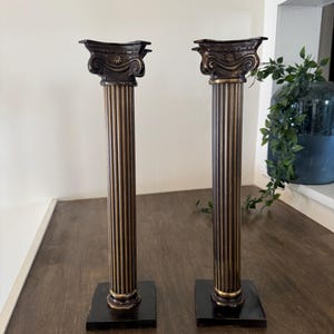 May include: Two bronze-coloured Corinthian-style columns. Each column has a square base and a fluted shaft with gold accents. The capitals are intricately detailed with scrollwork. The columns are displayed on a dark wooden surface.