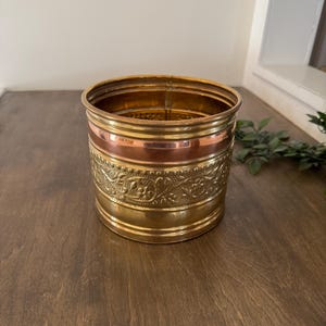 Könnte beinhalten: Ein zylindrischer Metallbehälter mit dekorativem Design. Der Behälter hat Bänder aus Gold, Kupfer und Messing. Das mittlere Band hat ein geprägtes Blumenmuster. Der Behälter steht auf einer Holzoberfläche.