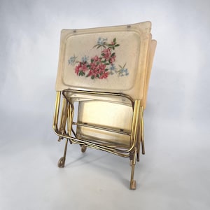 Può includere: Un set di quattro vassoi pieghevoli rettangolari con gambe in metallo dorato. Il vassoio superiore presenta una superficie color crema con una trama maculata e un'illustrazione floreale centrale di fiori rosa e blu con foglie verdi.