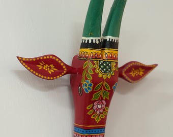 Pode incluir: Cabeça de touro de madeira pintada à mão, base vermelha vibrante. Chifres verdes com pontas pretas e padrões florais decorativos em amarelo, azul e rosa. Pequenos detalhes vermelhos em forma de asas em cada lado.