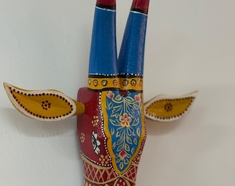Pode incluir: Escultura de cabeça de touro em madeira pintada à mão. A escultura apresenta um corpo vermelho com intrincados padrões dourados e azuis, chifres azuis e detalhes amarelos. O touro tem pequenas asas amarelas em ambos os lados.