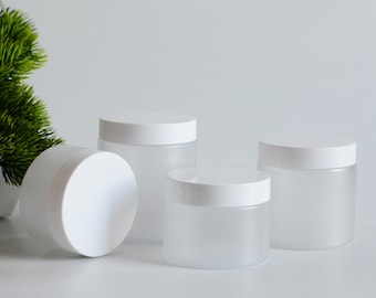 May include: Four frosted glass jars with white lids are arranged on a white surface. One jar is on its side, while the others stand upright. The jars are cylindrical and appear to be empty. The background is a plain white.