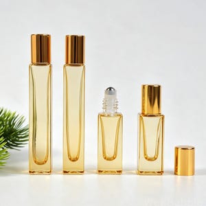 May include: Four rectangular glass roller bottles with gold caps. Two tall bottles are capped, one shorter bottle is uncapped showing a metal roller ball, and one short bottle is capped with its gold lid placed separately to the right side.