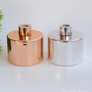 May include: Two metallic cylindrical containers, one copper-coloured and the other silver, with matching small, cylindrical tops. The containers are likely for home fragrance products, such as diffusers or candles. The background is white.