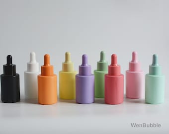 Puede incluir: Diez botellas de vidrio con gotero en varios colores pastel, incluyendo negro, blanco, naranja, amarillo, morado, verde, rosa y verde claro. Las botellas están dispuestas en una fila sobre una superficie blanca.