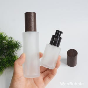 May include: Two frosted glass bottles with dark brown wood-grain caps. One bottle is tall and cylindrical with a wood-grain cap, while the other is shorter, rectangular, and features a black pump dispenser. The text WenBubble is visible.