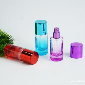 Op de afbeelding: Drie cilindrische glazen parfumflesjes met metalen doppen. Eén flesje is rood met een rode dop, één is blauw met een blauwe dop en één is paars met een paarse dop. Het paarse flesje is afgebeeld zonder dop, waardoor de verstuiver zichtbaar is.
