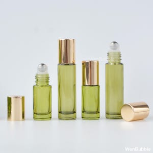 May include: Four cylindrical glass roller bottles with a light olive green tint. Two bottles are capped with shiny gold lids, while two are uncapped, revealing clear roller balls. The text WenBubble is visible in the background.