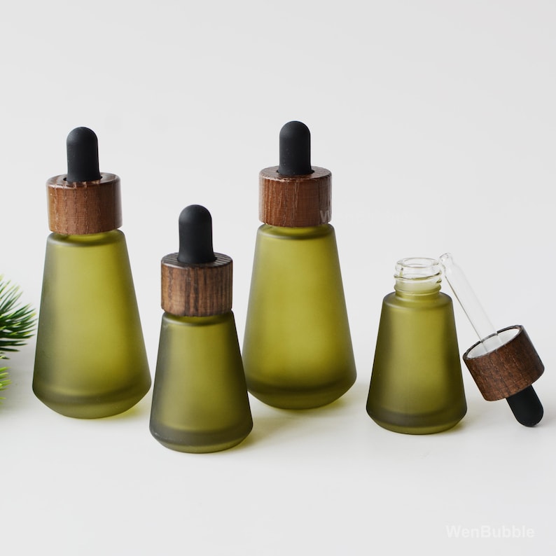 May include: Four olive green glass dropper bottles with wooden caps and black droppers. One bottle is open with the dropper and cap separate. The bottles are arranged on a white surface.
