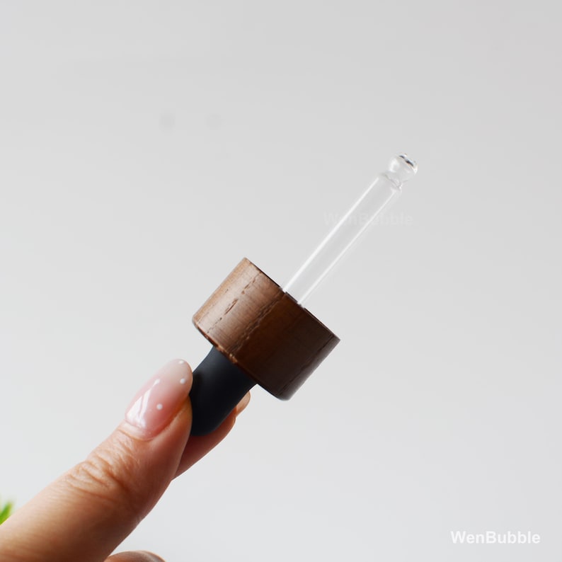 May include: A close-up of a clear glass dropper with a brown wooden cap and a black rubber bulb. The dropper is angled, with the tip pointing towards the upper right. The background is a soft, neutral white.