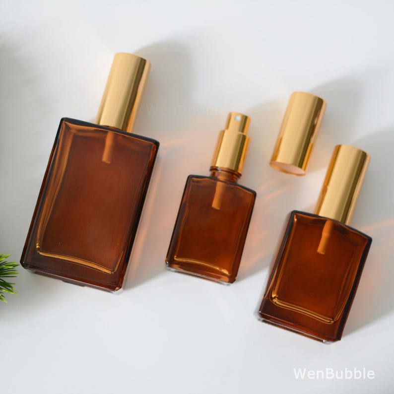 May include: Three amber glass bottles with gold atomisers. The bottles are different sizes and are arranged on a white surface.