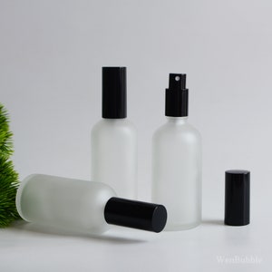 May include: Three frosted glass spray bottles with black caps. One bottle is lying on its side, while the other two stand upright. One of the upright bottles has a spray nozzle. The bottles are empty and ready to be filled.