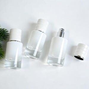 Op de afbeelding: Drie cilindrische glazen spuitflessen met witte doppen en één losse witte dop. De flessen hebben een heldere glazen basis en een wit ondoorzichtig bovendeel. Het zilverkleurige spuitmechanisme is zichtbaar op een van de flessen.
