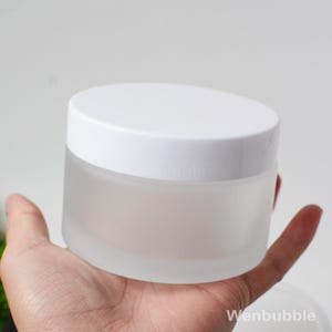 May include: A frosted glass jar with a white lid. The jar is round and held in a hand. The jar is empty and ready to be filled with product. The jar has a minimalist design.