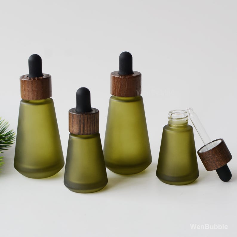 May include: Four olive green glass dropper bottles with wooden caps and black droppers. One bottle is open, with the dropper partially inserted. The bottles are arranged on a white surface.
