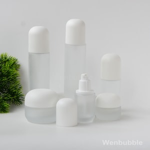 Peut inclure: Ensemble de contenants cosmétiques en verre dépoli avec des bouchons blancs arrondis. L'ensemble comprend des flacons, des pots et un distributeur à pompe, de tailles variées. Les contenants sont disposés sur une surface blanche.