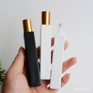 May include: Three rectangular perfume bottles with gold-coloured caps are held in a hand. One bottle is black, and two are white. One of the white bottles has a rollerball applicator.
