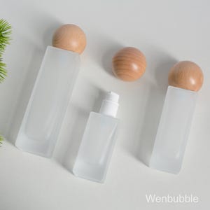 Peut inclure: Trois flacons en verre dépoli avec des bouchons en bois et un flacon à pompe blanc sont disposés sur une surface blanche. Les flacons sont rectangulaires et les bouchons en bois sont sphériques. Les flacons sont vides.