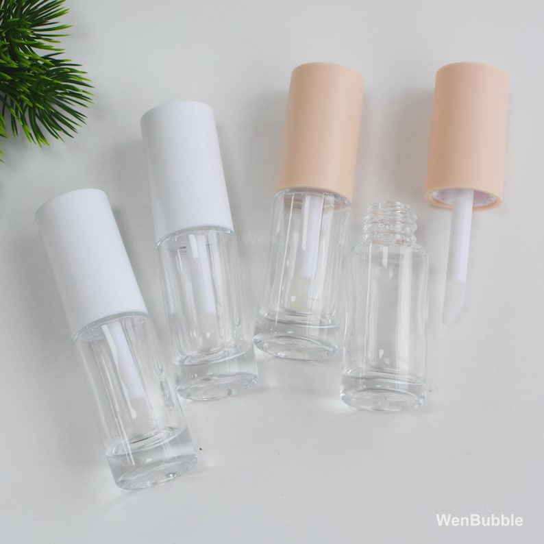 Peut inclure: Plusieurs tubes de gloss transparents avec des bouchons blancs et p&ecirc;che sont dispos&eacute;s sur une surface blanche. Un tube est ouvert, r&eacute;v&eacute;lant l'applicateur. Les flacons sont cylindriques et vides.