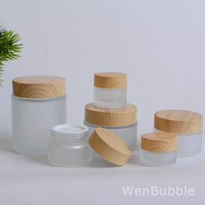 Könnte beinhalten: Eine Sammlung von Gläsern aus mattiertem Glas mit Holzdeckeln. Die Gläser sind in verschiedenen Größen und Formen erhältlich, einige mit sichtbarem Inhalt. Die Deckel sind hellbraun, und die Gläser sind auf einer weißen Oberfläche angeordnet. Der Text "WenBubble" ist sichtbar.