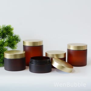 Könnte beinhalten: Eine Auswahl an braunen Glasbehältern mit goldenen Deckeln. Die Behälter sind in verschiedenen Größen erhältlich, einer ist geöffnet und zeigt das Innere. Sie sind auf einer weißen Oberfläche angeordnet, mit grünem Laub im Hintergrund.