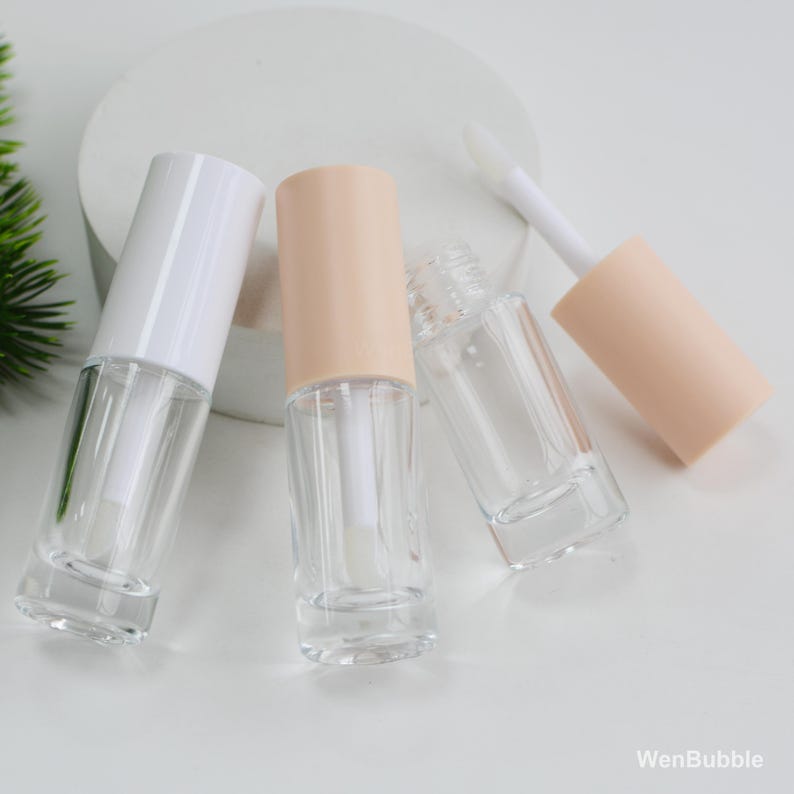 Peut inclure: Flacons de gloss transparents avec des bouchons blancs et p&ecirc;che. Un flacon est ouvert, r&eacute;v&eacute;lant la baguette applicatrice. Les flacons sont dispos&eacute;s sur une surface blanche, avec un soup&ccedil;on de verdure en arri&egrave;re-plan.