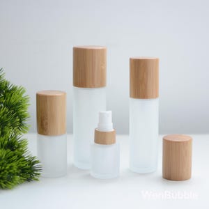 Puede incluir: Colección de botellas y frascos de vidrio esmerilado con tapas de bambú natural. El conjunto incluye varios tamaños, desde pequeños pulverizadores hasta recipientes cilíndricos más grandes, todos con una estética minimalista.
