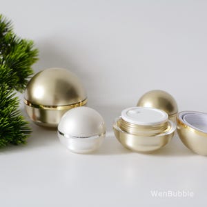 Peut inclure: Quatre pots cosmétiques ronds dorés et blancs avec des couvercles. Les pots sont disposés sur une surface blanche. Les pots ont une finition lisse et brillante.