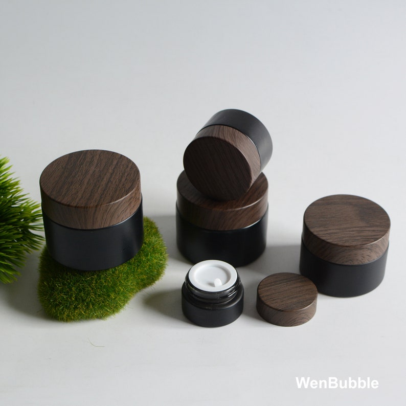 May include: Set of five black glass jars with wooden lids. The jars are different sizes and are stacked on top of each other. The jars are on a white surface.