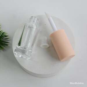 Peut inclure: Un flacon de gloss transparent vide avec un applicateur p&ecirc;che et une baguette blanche. Le flacon est &agrave; c&ocirc;t&eacute; d'un bouchon transparent. Les articles sont sur une surface blanche et circulaire. La marque "WenBubble" est visible.