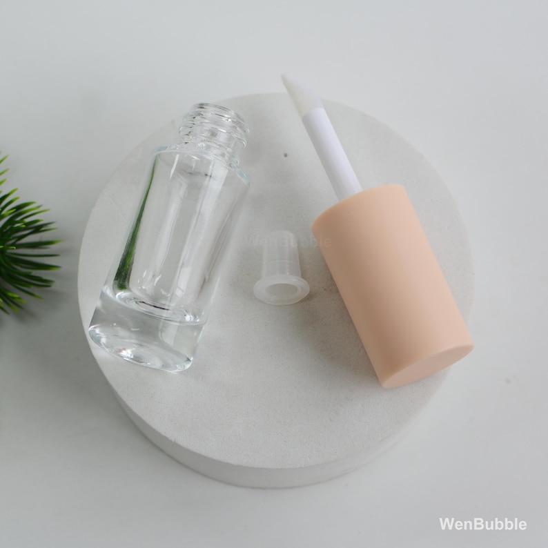 Peut inclure: Un flacon de gloss transparent vide avec un applicateur p&ecirc;che et une baguette blanche. Le flacon est &agrave; c&ocirc;t&eacute; d'un bouchon transparent. Les articles sont sur une surface blanche et circulaire. La marque "WenBubble" est visible.