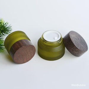 Op de afbeelding: Twee olijfgroene glazen potten met houten deksels. Eén pot is open en onthult een wit interieur. De deksels hebben een donker houtnerfpatroon. De potten staan op een witte ondergrond.