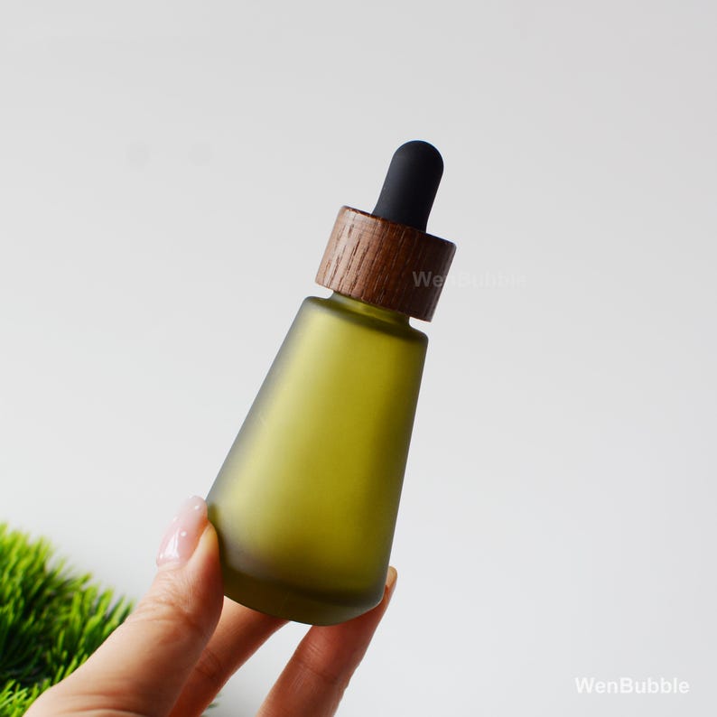 May include: A green glass essential oil bottle with a wooden cap and black dropper. The bottle has a matte finish and a conical shape. The wooden cap has a dark brown color.