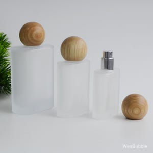 Puede incluir: Tres frascos de perfume de vidrio esmerilado con tapones esféricos de madera. Los frascos varían en tamaño y forma, uno de ellos con un pulverizador plateado. Los tapones de madera son de color marrón claro y los frascos son blancos.