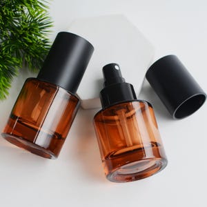 May include: Two amber glass spray bottles with black spray tops and black caps. The bottles are rectangular with rounded corners. One bottle has the cap on, and the other has the cap off. The background is white with a green plant in the upper left corner.