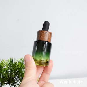May include: A small, clear glass bottle with a green gradient, a wooden cap, and a black dropper. The bottle is held in a hand, with green foliage in the background. The bottle is labeled with the text "Wenbubble."