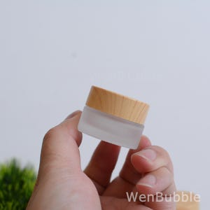 May include: A small, frosted glass jar with a light wood lid. The jar is square with rounded corners. The lid is a light, natural wood color. The jar is held in a person's hand. The text "WenBubble" is visible in the bottom right corner.