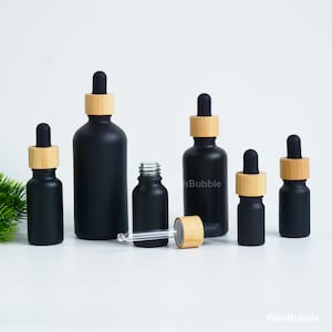 May include: A collection of matte black glass dropper bottles of varying sizes, each featuring a light-coloured wooden collar and a black rubber bulb. One bottle is shown open with its glass pipette detached and lying horizontally in front.
