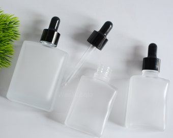 May include: Three frosted glass bottles with black dropper tops. The bottles are rectangular and have a frosted finish. The bottles are on a white surface.